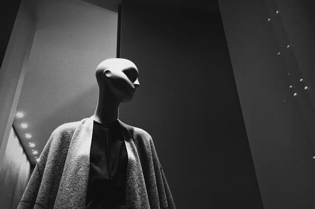 A close-up shot of a disturbing mannequin in a dark and dusty room in a video game. The mannequin's face is partially obscured by shadows, and there are cobwebs and dust particles floating in the air. The scene conveys a sense of decay and unease.